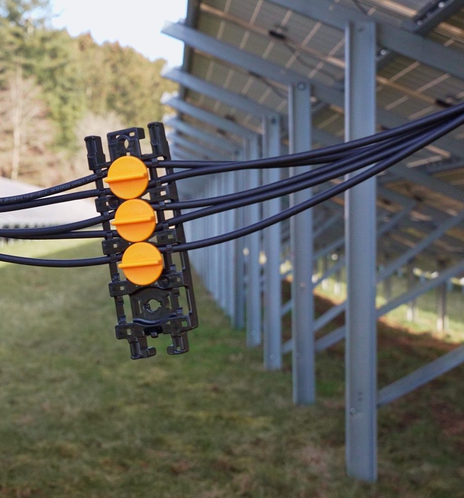 ENGESOLAR Clip mit drei verriegelten Deckeln und eingelegten Solarleitungen vor einer Freiflächenanlage
